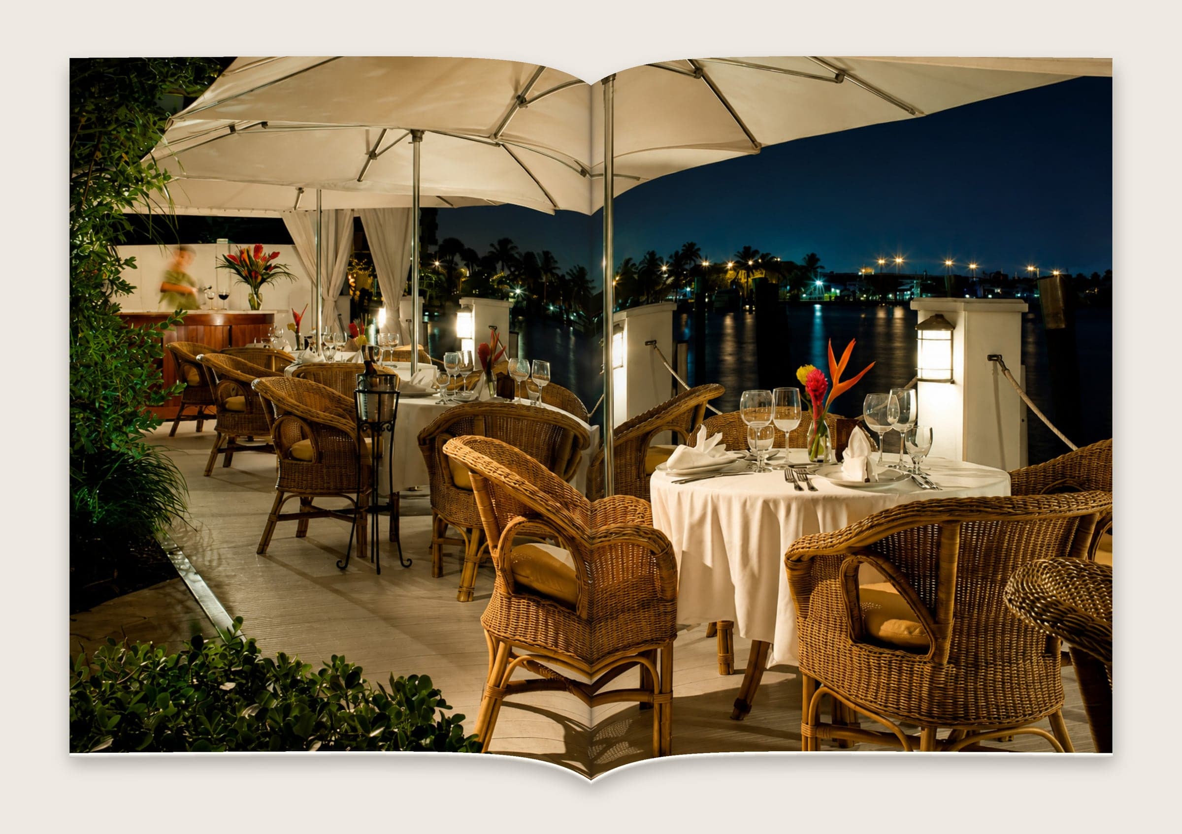 Photograph across a spread of wicker chairs and set tables under umbrellas beside the water at dusk.